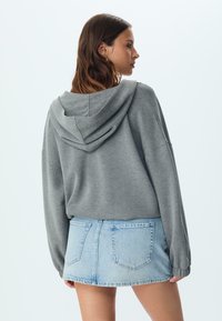 Šedá skrátená mikina s kapucňou, vo voľnom strihu a dlhými rukávmi, skombinovaná s krátkou džínsovou sukňou z ľahkého denimu s viditeľnými zadnými vreckami.