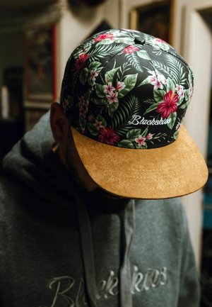Gorra con estampado floral y parte superior de tela negra que presenta diseños en rosa y verde, y una visera de gamuza beige. Bordado "Blackskies" en la parte frontal.