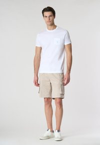 T-shirt bianco in cotone con dettagli della tasca blu chiaro, abbinato a pantaloni cargo beige con più tasche laterali, e sneakers bianche.