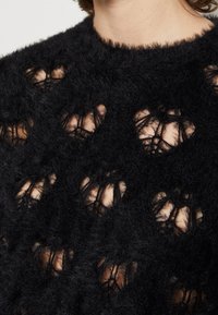 Pull en tricot noir avec une finition texturée et duveteuse, présentant un motif ajouré qui crée des découpes en forme de cœur tout au long.