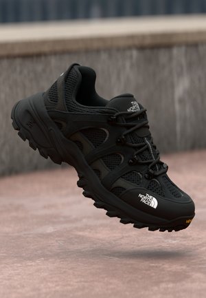 Zwarte sportieve schoen met een mesh bovenwerk, versterkte accenten, dikke zool met grip en een opvallend logo aan de zijkant.