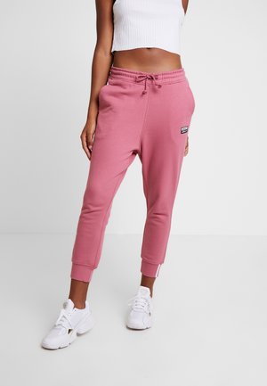 Kobieta w różowych spodniach dresowych Adidas, białym krótkim prążkowanym topie i białych trampkach, stoi na jednolitym jasnym tle.