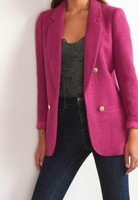 Blazer texturé fuchsia avec un col cranté, des boutons dorés et des accents roses aux poignets, porté avec un caraco en dentelle noire et un jean foncé.