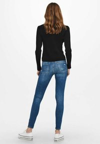 Jeans de mezclilla azul ajustados con un lavado descolorido, bolsillos traseros y detalles desgastados, combinados con una camiseta negra de manga larga y zapatillas blancas.