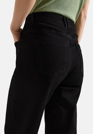 Personne portant un jean noir avec des poches arrière visibles et les mains partiellement dans les poches, assorti d'un haut vert clair.