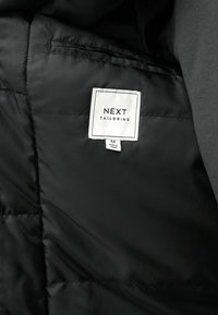 Intérieur d'une doudoune noire avec une étiquette blanche : "NEXT TAILORING," taille M, avec une texture lisse et une doublure douce.