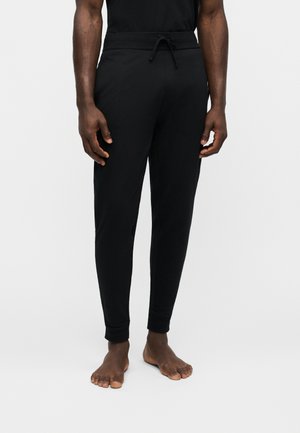 Homme portant un pantalon de jogging noir à cordon, debout pieds nus sur un fond clair uni.