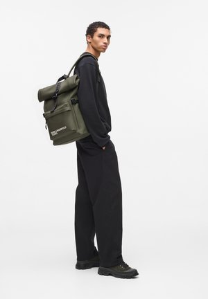 Rucsac verde cu capac rulabil, curele negre, buzunar frontal și detaliu cu logo. Modelul poartă un pulover negru și pantaloni largi, evidențiind funcționalitatea.