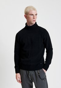 Maglione nero a collo alto in maglia con polsini e orlo a coste. Presenta una finitura texturizzata e una vestibilità morbida. Abbinato a pantaloni grigi.