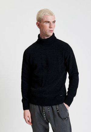 Maglione nero a collo alto in maglia con polsini e orlo a coste. Presenta una finitura texturizzata e una vestibilità morbida. Abbinato a pantaloni grigi.