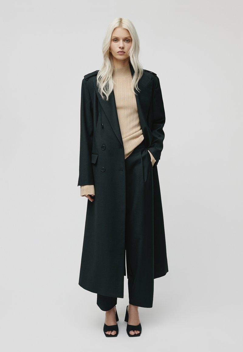 Donkergroene dubbelzijdige jas met een gestructureerd ontwerp, gedragen over een geribbeld beige top, gecombineerd met wijde zwarte broek en zwarte hakken.