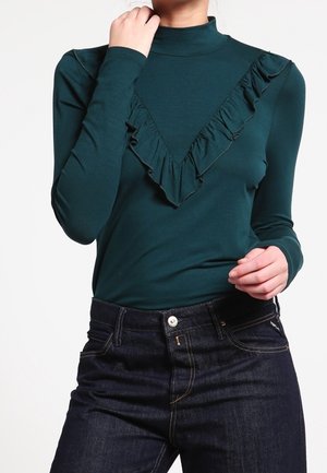 Vrouw draagt een donkergroene top met lange mouwen en een gerimpeld V-vormig detail, gecombineerd met donkere denim jeans, tegen een effen achtergrond.