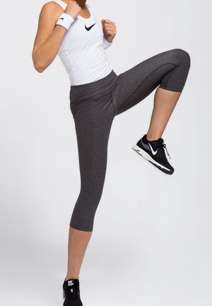 Person iført hvid tanktop og grå leggings, løfter knæet, iført sorte Nike-sneakers og hvide håndledsbind, mod ensfarvet baggrund.