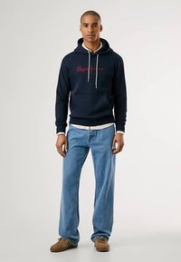 Sweatshirt azul-marinho com bolso canguru frontal e cordões brancos, com um logótipo vermelho. Combinado com jeans azuis claros de perna reta e sapatos castanhos.