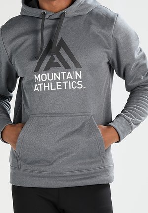 Person iført en grå hættetrøje med et bjergformet logo og teksten "Mountain Athletics", hænderne i frontlommen, sorte bukser synlige.