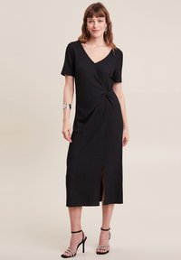 Schwarzes Midi-Kleid mit V-Ausschnitt, kurzen Ärmeln, gedrehtem Knoten-Detail an der Taille und einem seitlichen Schlitz. Texturiertes Material sorgt für visuelles Interesse.