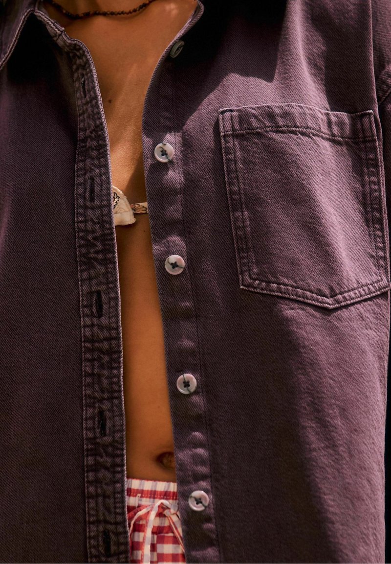 Chemise violette déboutonnée sur un torse nu avec un collier visible, bretelle de haut de bikini et short à cordon à carreaux rouge et blanc.