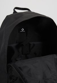 Intérieur d'un sac à dos en toile noire avec une texture lisse, arborant une étiquette Converse et une sangle sécurisée pour l'organisation.
