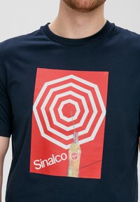 Navy blauwe T-shirt met een grote rode vierkante grafiek met concentrische witte hexagonen en een fles Sinalco frisdrank onderaan.