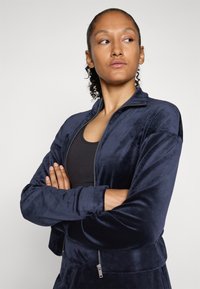 Žena s kučeravými vlasmi v námornícky modrej zipsovej sametovej bunde a čiernom topu, s prekríženými rukami, pozerajúca sa mierne na stranu proti jednoduchému pozadiu.