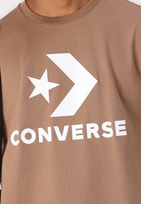 Ruisin värinen puuvillat-paita, jossa on suuri valkoinen tähti, nuoli grafiikka ja rohkea "CONVERSE" -teksti alareunassa. Pyöreä kaula-aukko.