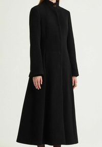 Cappotto lungo nero in tessuto strutturato, con colletto alto, chiusura con bottoni e silhouette svasata, che arriva sotto il ginocchio.