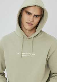 Hoodie vert clair en tissu doux, avec un imprimé sur le devant qui indique "JE SUIS UNE PERSONNE WEEK-END" en texte blanc. Cordon de serrage à la capuche.