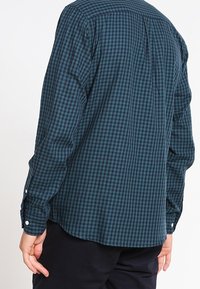 Herenoverhemd met lange mouwen in donker turquoise met een geruit patroon. Voorzien van knoopmanchetten en een schouderstuk aan de achterkant. Gemaakt van een katoenmengsel.