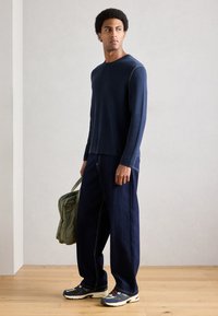 Tumši zils adīts krekls ar garām piedurknēm un gaišu šūšanu, kombinēts ar tumšām džinsa biksēm ar platām kājsaitēm un zaļu somu, pabeigts ar krāsainām sportiskām kedām.