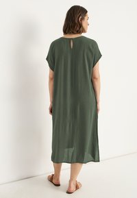 Langes, wehendes grünes Kleid mit kurzen Ärmeln, das ein Schlüssel-Detail am Rücken und einen strukturierten Stoff aufweist. Flache Sandalen vervollständigen den Look.