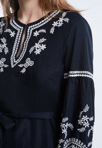 Blouse noire à manches longues avec broderie florale blanche sur la poitrine, les manches et le col, dotée d'une ouverture en forme de clé et d'une ceinture nouée à la taille.