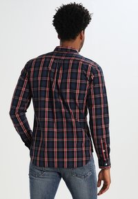 Camicia a quadri blu navy con dettagli rossi e bianchi, maniche lunghe, polsini con bottone e design dritto sul retro, realizzata in tessuto intrecciato.