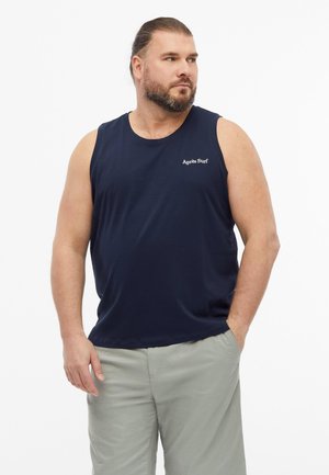 Mężczyzna z brodą i związanymi do tyłu włosami, ubrany w granatową bezrękawnik z napisem „Après Surf” i jasnoszorty, patrzący na bok.