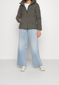 Oliivinvihreä toppatakki hupulla, vetoketjulla ja kahdella etutaskulla, yhdistettynä vaaleansinisiin leveälahkeisiin farkkuihin ja urheilullisiin kenkiin.