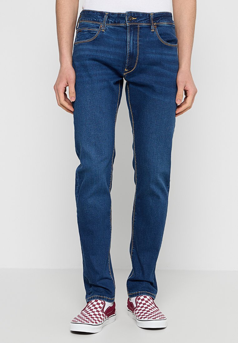 Schmale blaue Jeans aus Denim mit goldenen Nähten und fünf Taschen. Getragen mit rot-weiß karierten Slippern.