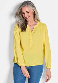 Blusa gialla a maniche lunghe con scollo a V, quattro bottoni sul davanti e dettaglio con bottoni sui polsini. Realizzata in tessuto leggero, con una texture liscia.