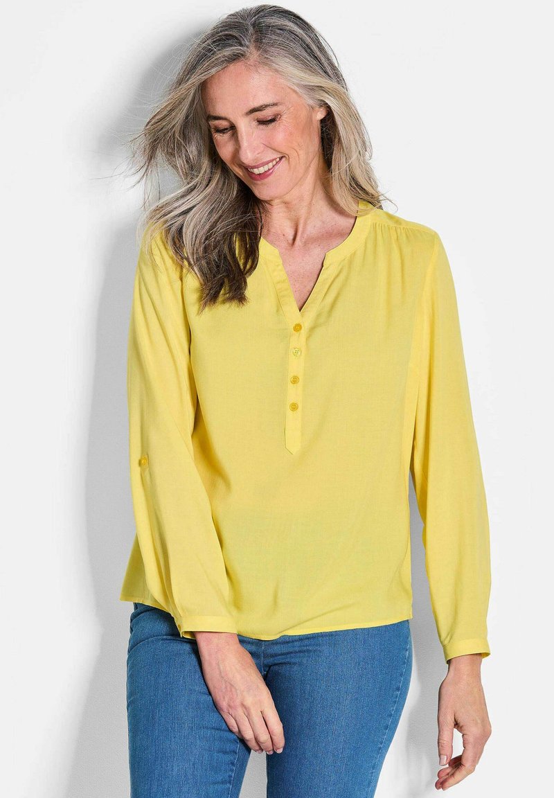 Blusa gialla a maniche lunghe con scollo a V, quattro bottoni sul davanti e dettaglio con bottoni sui polsini. Realizzata in tessuto leggero, con una texture liscia.