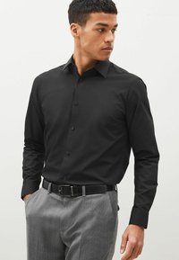 Camisa negra de botones con mangas largas, diseño ajustado y textura suave; combinada con pantalones texturizados gris claro y un cinturón negro.