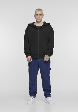 Młoda osoba o krótkich blond włosach stoi twarzą do przodu, ubrana w czarną bluzę na zamek, luźne niebieskie dżinsy i białe trampki.