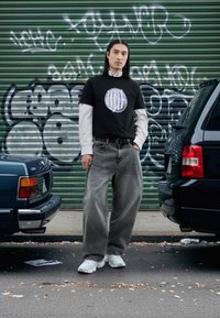 Must t-särk, millel on ümmargune graafika, kandes seda valge triibulise pika varrukaga särkide kohal. Hallid laiade säärtega teksad ja valged vabaajajalanõud viimistlevad välimuse.