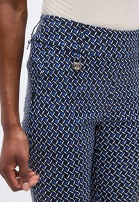 Κοντινό πλάνο ενός ατόμου που φοράει ναυτικό μπλε παντελόνι με γεωμετρικό μπλε και λευκό μοτίβο και ένα μικρό ασημένιο φερμουάρ στη μέση.