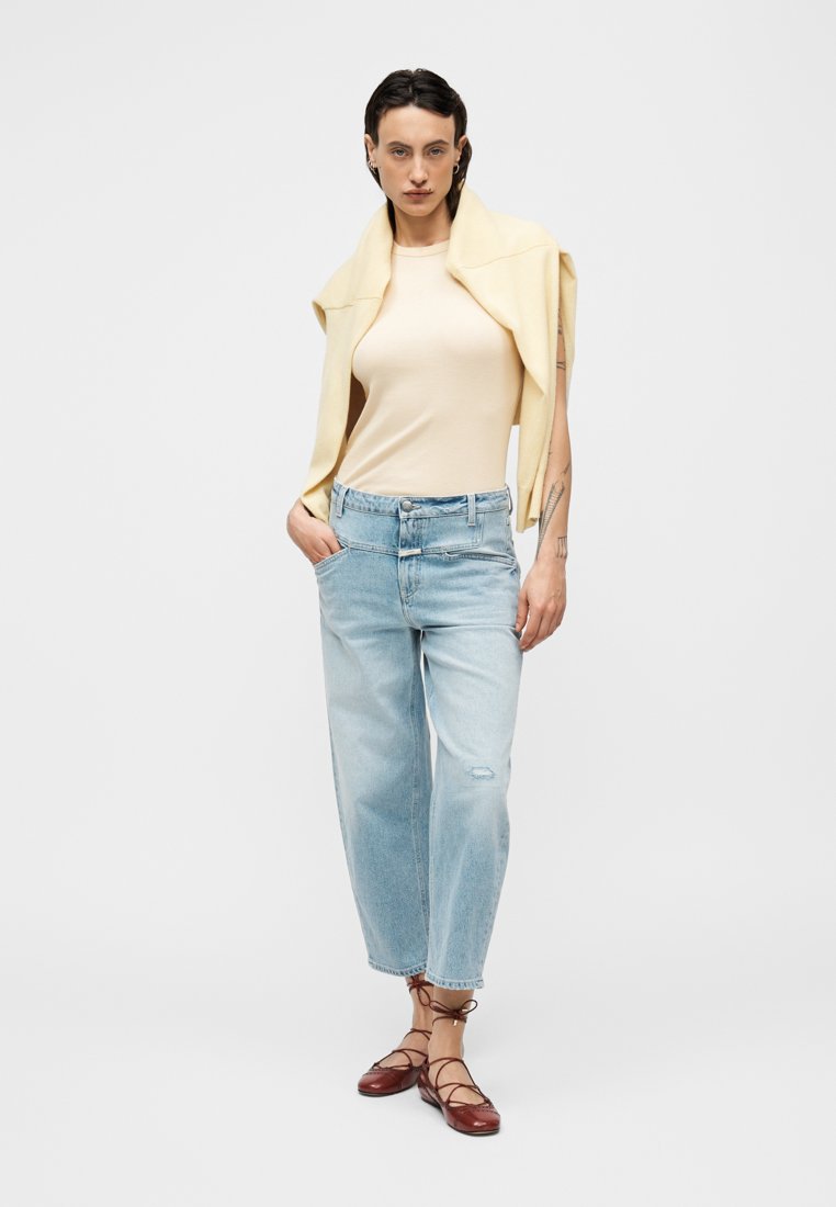 Haut beige ajusté, jean bleu clair décontracté avec une finition délavée et chaussures marron à lacets. Un pull jaune pâle drapé sur les épaules.