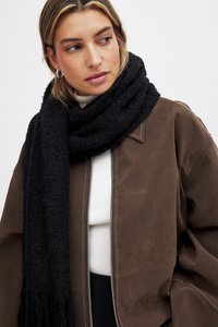 Giacca in finta pelle scamosciata marrone con zip, dolcevita bianco a coste e una sciarpa nera di maglia spessa. Texture che varia tra liscio e peloso.