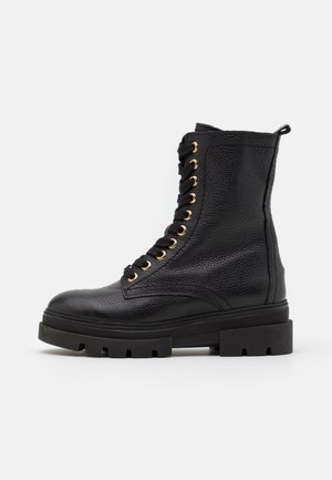 Botte de combat en cuir noir texturé à lacets avec semelle épaisse à crampons et œillets dorés, vue de profil sur fond blanc.
