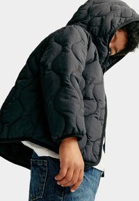 Giacca invernale nera trapuntata con cappuccio, caratterizzata da una superficie testurizzata e polsini elasticizzati. Indossata sopra una camicia bianca e jeans in denim.