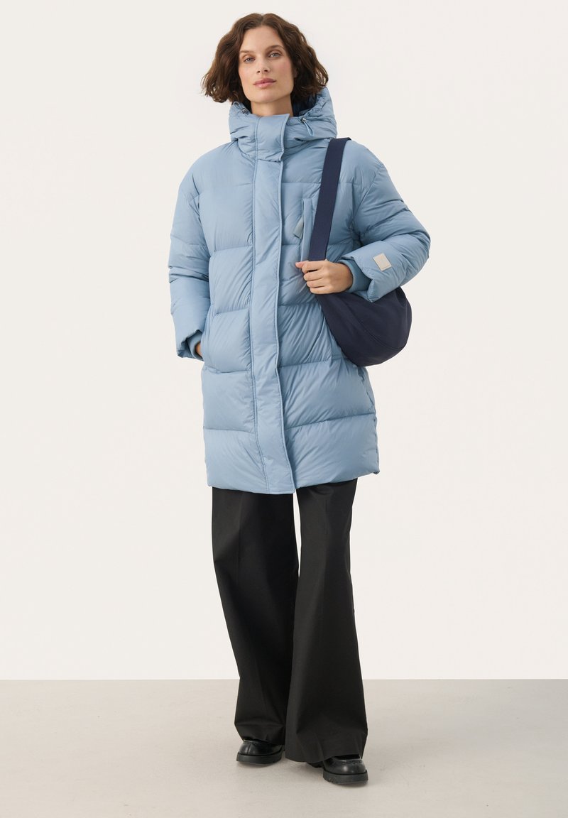 Hellblauer Puffercoat mit hohem Kragen, Frontreißverschluss und Nahtdetails. Kombiniert mit schwarzen weit geschnittenen Hosen und einer dunklen Tasche. Glatte Textur.