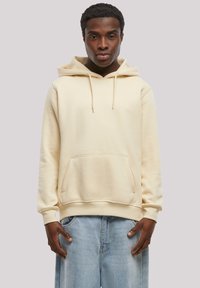 Cream hoodie made of soft fabric with a kangaroo pocket, featuring a drawstring hood and ribbed cuffs, paired with light blue jeans.