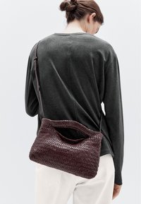 Borsa in pelle intrecciata di un profondo bordeaux, con manico curvo e tracolla regolabile, caratterizzata da una texture liscia e da una forma morbida.