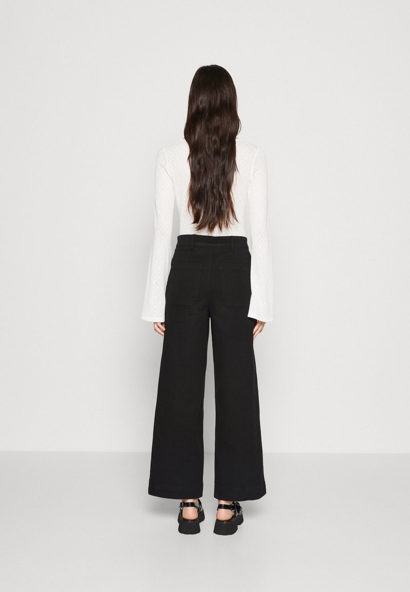 Žena stojící zády, oblečená v bílém topu s dlouhým rukávem a rozšířenými manžetami, černých širokých kalhotách a černých platformových sandálech.