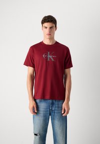 Bordo pamučna majica s okruglim izrezom, kratkim rukavima i siva logotip ispisana "Calvin Klein" te velikim "K" na prednjoj strani.